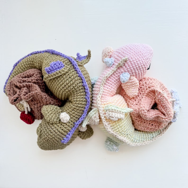 K&ouml;nnte beinhalten: Zwei geh&auml;kelte Stoffdrachen, einer gr&uuml;n und einer rosa, mit wei&szlig;en Akzenten. Die Drachen sind aneinander gekuschelt.