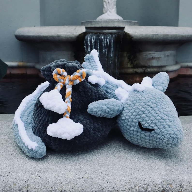 K&ouml;nnte beinhalten: Zwei geh&auml;kelte Stofftiere, ein grauer Drache mit wei&szlig;em Bauch und eine schwarze Tasche mit einem wei&szlig;-orangenen Band.