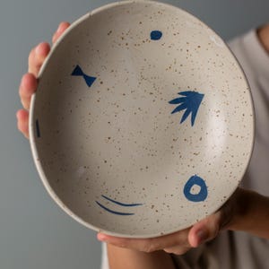 Puede incluir: Un cuenco de cerámica moteada con una base de color crema y diseños abstractos azules. El cuenco presenta una variedad de formas azules, incluyendo puntos, una pajarita y un diseño en forma de hoja. El cuenco es sostenido por una persona.