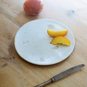 May include: A white ceramic plate with a speckled finish, featuring two slices of peach and a red, speckled egg on a wooden table. A silver butter knife is also on the table.