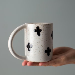 Può includere: Una tazza di ceramica bianca con finitura maculata e disegni a croce neri. La tazza ha un manico grande e curvo ed è tenuta da una mano. La tazza è di colore neutro e adatta per caffè, tè o altre bevande.