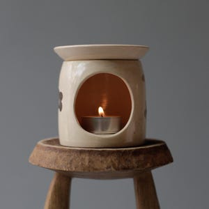 May include: A cream-colored ceramic essential oil diffuser with a small tea light candle burning inside. The diffuser has a round opening and is decorated with dark brown floral accents. It sits on a small, rustic wooden stool.