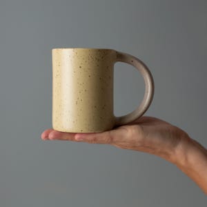 Op de afbeelding: Een beige keramische mok met een afgeronde handgreep en een cilindrische vorm. De mok wordt in een hand gehouden tegen een grijze achtergrond. De buitenkant van de mok heeft kleine, donkerbruine spikkels.