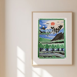 May include: A framed print of the cover art for the book "The Hobbit" by J.R.R. Tolkien. The illustration depicts a landscape with mountains, a lake, and trees in shades of blue, green, and brown.