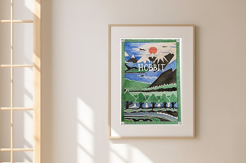 May include: A framed print of the cover art for the book "The Hobbit" by J.R.R. Tolkien. The illustration depicts a landscape with mountains, a lake, and trees in shades of blue, green, and brown.