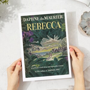 May include: A book cover with a green and blue illustration of a house and a couple walking on a path. The title "REBECCA" is in large yellow letters and the author "DAPHNE du MAURIER" is in smaller black letters. The text "A brilliant novel of an unforgettable woman. by the author of JAMAICA INN" is at the bottom of the cover.