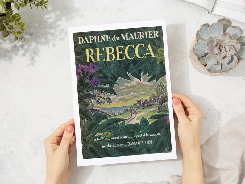 May include: A book cover with a green and blue illustration of a house and a couple walking on a path. The title "REBECCA" is in large yellow letters and the author "DAPHNE du MAURIER" is in smaller black letters. The text "A brilliant novel of an unforgettable woman. by the author of JAMAICA INN" is at the bottom of the cover.