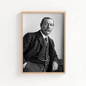May include: Black and white framed portrait of a man in a suit and tie. The man has a mustache and is leaning against a dark surface. The portrait is in a light brown wooden frame and is displayed against a white wall.