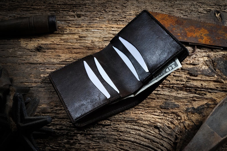 May include: A brown leather wallet with a billfold and card slots, open to reveal a &pound;10 note. The wallet is lying on a rustic wooden surface.