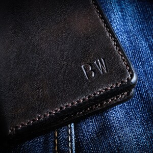 May include: A black leather wallet with a stitched edge and the initials "BW" embossed on the front. The wallet is lying on a blue denim background.
