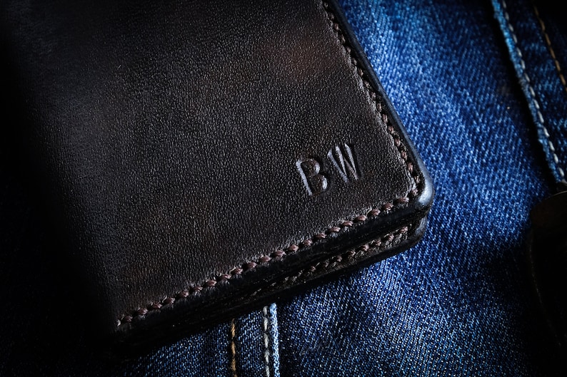 May include: A black leather wallet with a stitched edge and the initials "BW" embossed on the front. The wallet is lying on a blue denim background.