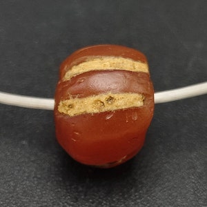 Könnte beinhalten: Nahaufnahme einer roten Karneolperle mit drei goldenen Streifen. Die Perle befindet sich auf einer weißen Schnur.