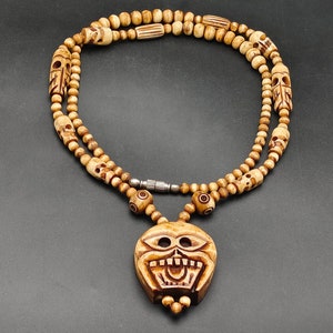 Puede incluir: Un collar de madera marrón con un colgante de calavera. El collar está hecho de múltiples hilos de cuentas con detalles de calavera tallados. El colgante es una calavera grande y detallada con una sonrisa de dientes.