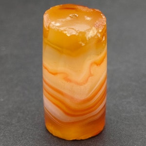 May include: A cylindrical agate bead with a banded pattern of orange, white, and brown. The bead has a smooth, polished surface.