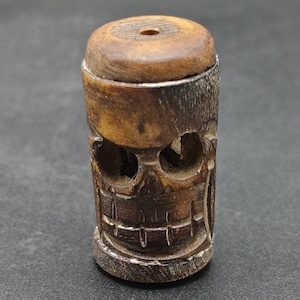 Può includere: Un piccolo perno di legno a forma di teschio con intagli intricati. Il perno è marrone e ha un foro nella parte superiore per la corda.