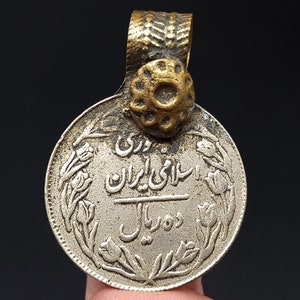 Puede incluir: Colgante de moneda de plata antigua con una fianza dorada. La moneda presenta escritura persa y diseños florales, con una inscripción central y la denominación "2 Rial". El colgante tiene una fianza decorativa y texturizada.