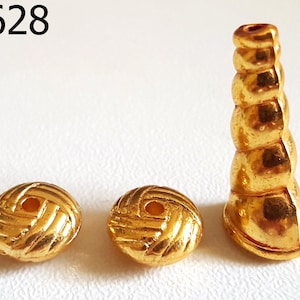 Könnte beinhalten: Drei goldfarbene Perlen und eine goldfarbene zylindrische Perle. Die beiden runden Perlen haben ein gewebtes Muster. Die zylindrische Perle ist mit abgerundeten Segmenten gestapelt. Der Text "UK628" befindet sich oben links.