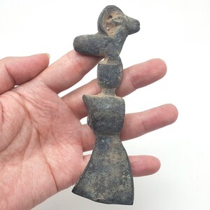 May include: A dark grey stone sculpture of a ram with a long, narrow head and horns. The sculpture is in the shape of a stylised human figure with a wide, flat base.