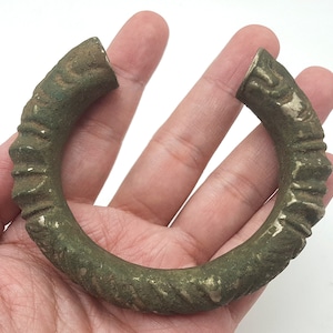 Puede incluir: Un brazalete de metal verde con un diseño texturizado. El brazalete está abierto y tiene una forma redondeada.