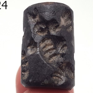 Könnte beinhalten: Ein schwarzer zylindrischer Stempel mit aufwendigen Schnitzereien. Der Stempel hat eine raue, strukturierte Oberfläche und scheint aus Stein zu sein.