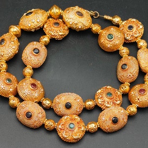 May include: A gold necklace with multiple oval-shaped pendants, each with a different colored gemstone in the center. The necklace is made of gold beads and has a delicate, intricate design.