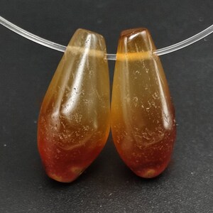 Puede incluir: Dos cuentas de cornalina en forma de lágrima con un color naranja y rojo translúcido. Las cuentas están enhebradas en un cordón transparente.