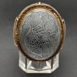 May include: A silver ring with an oval-shaped stone set in a gold-toned band. The stone is engraved with intricate Arabic calligraphy.