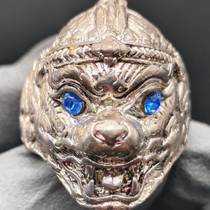 Peut inclure: Une bague en argent avec un design détaillé d'un visage de singe. Le singe a les yeux bleus et une expression féroce. La bague est une pièce unique et ferait un cadeau original.