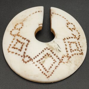 Könnte beinhalten: Ein weißer, runder Anhänger mit einem Loch in der Mitte. Der Anhänger ist mit einem braunen, geometrischen Muster verziert.