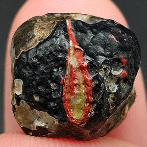 Puede incluir: Una cuenta de vidrio negra y roja con una franja blanca. La cuenta tiene una superficie rugosa y texturizada.