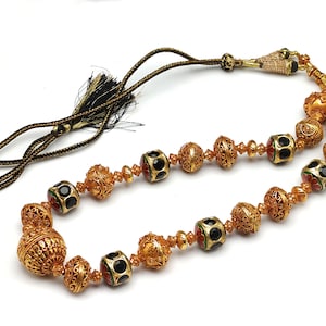 Peut inclure: Un collier en or avec des perles noires et rouges. Le collier a un pompon à l'extrémité.