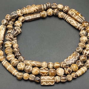 Puede incluir: Un collar largo hecho de cuentas marrones y blancas con patrones. Las cuentas están hechas de hueso y tienen un aspecto natural y rústico.