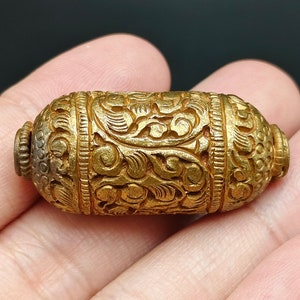 Puede incluir: Una cuenta cilíndrica de color dorado, tallada de forma intrincada con un diseño floral. La cuenta mide aproximadamente 5 cm de largo y tiene un acabado liso y pulido.