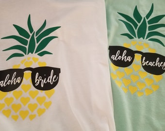 May include: Two white t-shirts with a pineapple design. The pineapple is wearing sunglasses and has the text "aloha bride" on one shirt and "aloha beaches" on the other.