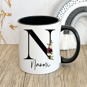 May include: White ceramic mug with a black rim and handle. The mug features a black letter "N" with floral accents and the name "Naomi" printed below.