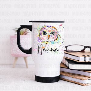 May include: White travel mug with a black handle and lid. The mug features a colourful watercolour painting of an owl with the word "Nanna" written below it.