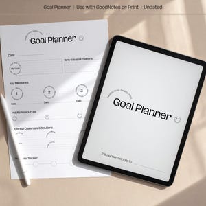 Könnte beinhalten: Ein digitales Tablet und ein gedruckter Zielplaner werden auf einer hellen Oberfläche gezeigt. Der Tablet-Bildschirm und die gedruckte Seite zeigen beide den Text "Goal Planner" in einer modernen Schriftart. Ein weißer Stift liegt auf dem Papier.