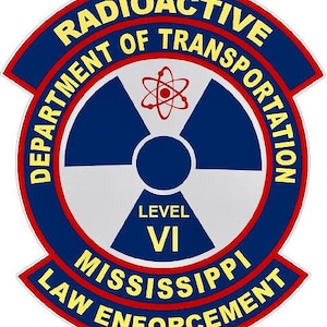 Pode incluir: Um adesivo circular azul e branco com o texto "Department of Transportation Radioactive" em letras amarelas. O adesivo também apresenta um símbolo de radiação estilizado e o texto "Level VI Mississippi Law Enforcement" em letras vermelhas e amarelas.
