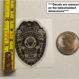 May include: A silver and black security enforcement officer decal with the words "Security Enforcement Officer SEO" printed on it. The decal is next to a US quarter for size comparison.