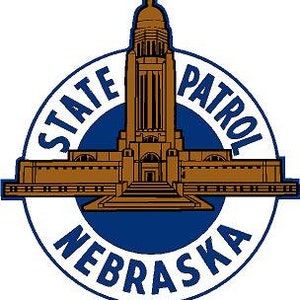 Op de afbeelding: Een blauw en gouden cirkelvormig embleem met de woorden "State Patrol Nebraska" rondom. Het midden van het embleem toont een bruine omtrek van het Nebraska State Capitol gebouw.
