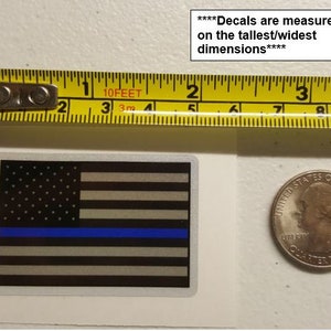 May include: A thin blue line American flag decal with a white background. The decal is measured on the tallest/widest dimensions. The decal is next to a ruler and a US quarter.
