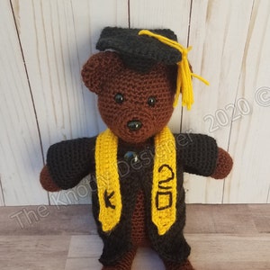May include: A brown crocheted teddy bear wearing a black graduation cap and gown with a yellow graduation sash. The sash has the year 2020 embroidered on it.