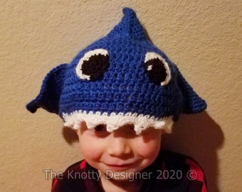 Puede incluir: Un gorro de tiburón de crochet azul con dientes blancos y ojos negros. El gorro lo lleva puesto un niño con una camisa de cuadros rojos y negros.
