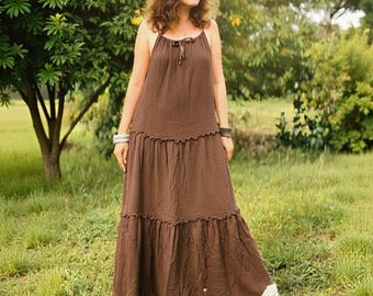 May include: A woman wearing a brown maxi dress with spaghetti straps and a tiered skirt, standing in a grassy field. She wears a floral crown and white boots. The dress has a relaxed fit, suitable for casual wear.