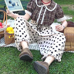 May include: A brown plaid short-sleeved blouse with a white lace-trimmed collar and cuffs, paired with cream-colored wide-leg trousers featuring a black polka dot pattern and ruffled hems, worn with a beige cap and dark-rimmed sunglasses.