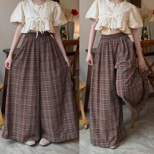 May include: A cream-coloured short-sleeved blouse with a ruffled neckline, button-down front, and a drawstring waist, paired with high-waisted, wide-leg trousers featuring a brown, black, and white plaid pattern, worn with a brown plaid beret.