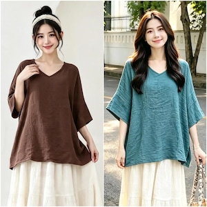 Pode incluir: Um conjunto de duas blusas largas com decote em V, mangas curtas e largas, e um acabamento de tecido texturizado e enrugado. Uma blusa é castanho-escura sólida e a outra é azul-petróleo. Ambas são combinadas com uma saia longa branca em camadas.