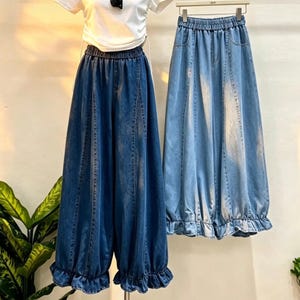 Puede incluir: Dos pantalones vaqueros anchos con cinturillas elásticas y dobladillos con volantes. Un par es de color azul índigo oscuro, mientras que el otro tiene un lavado azul más claro y desteñido. Ambos presentan costuras verticales frontales.