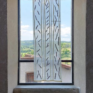 以下が含まれることがあります： 垂直な線と濃い茶色の葉のような形がパターン化された、透明感のある白いカーテン。カーテンは、緑の風景と青い空が見える窓に吊るされています。