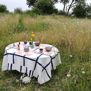Peut inclure: Une nappe blanche à carreaux bleus et roses est posée sur une table dans un champ d'herbe haute. La table est dressée avec une variété de petits bols et vases en céramique, dont certains sont remplis de fleurs.